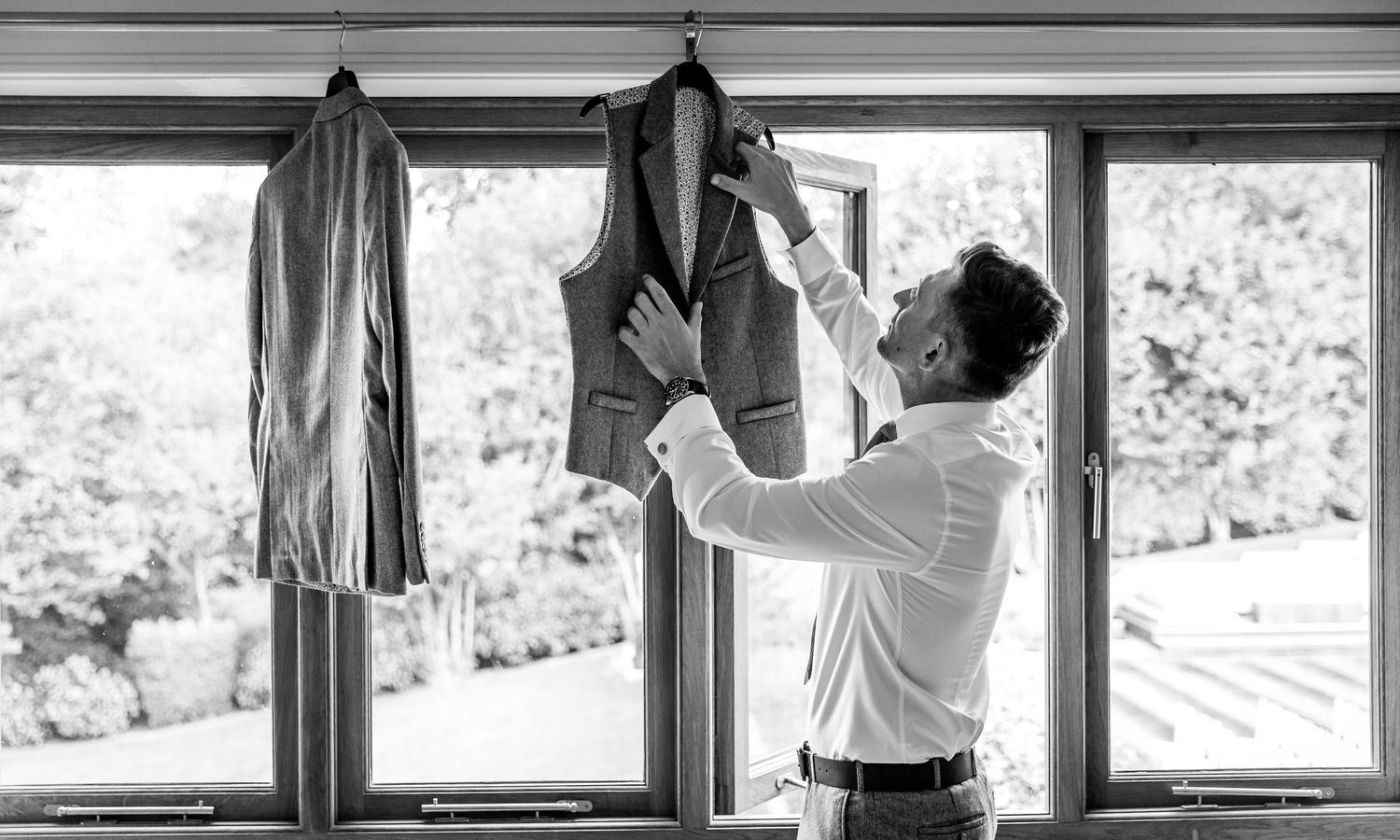 A person in a white shirt hangs dress shirts on a clothing rack by a bright sunlit window.