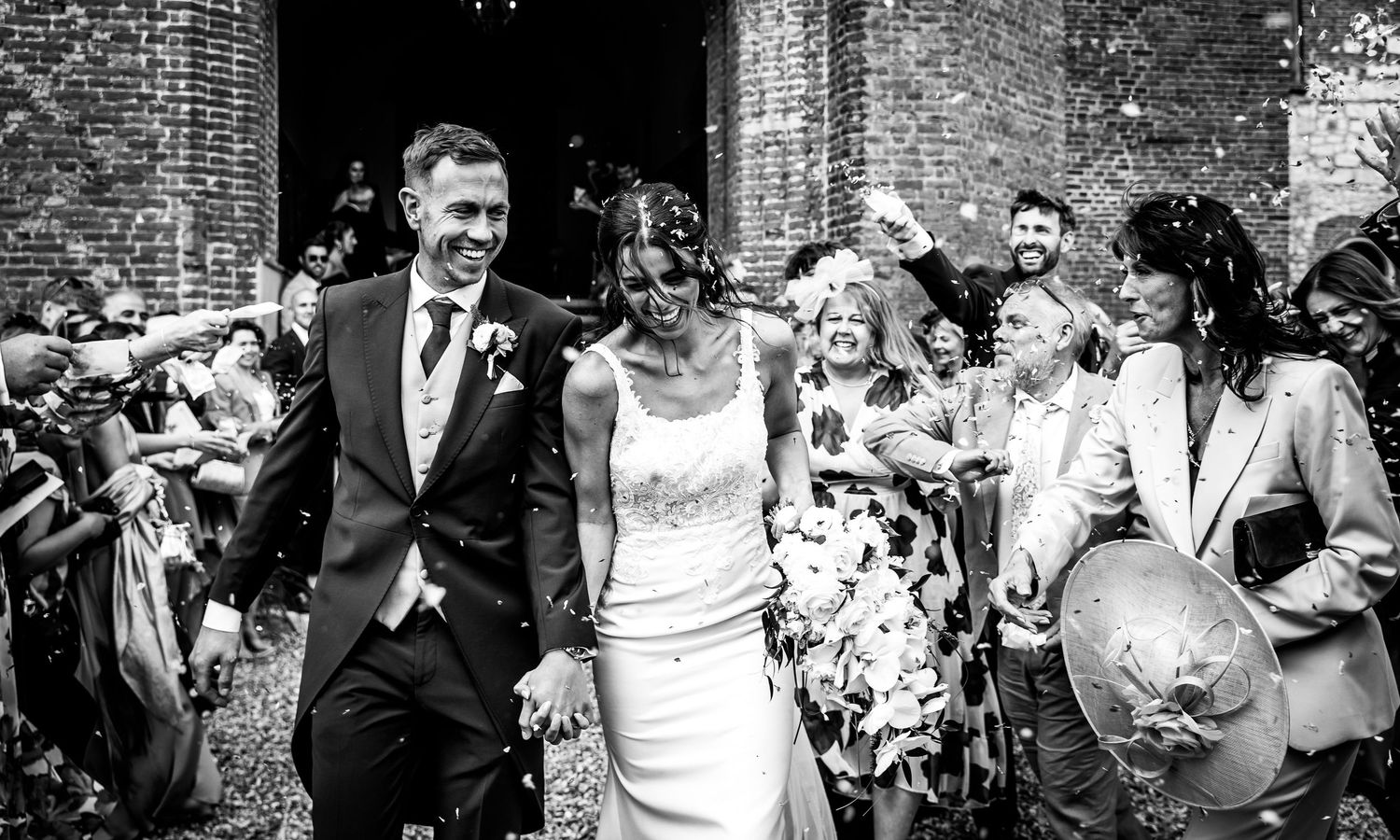 Wedding guests shower newlyweds with confetti outside a brick church in black and white photography.