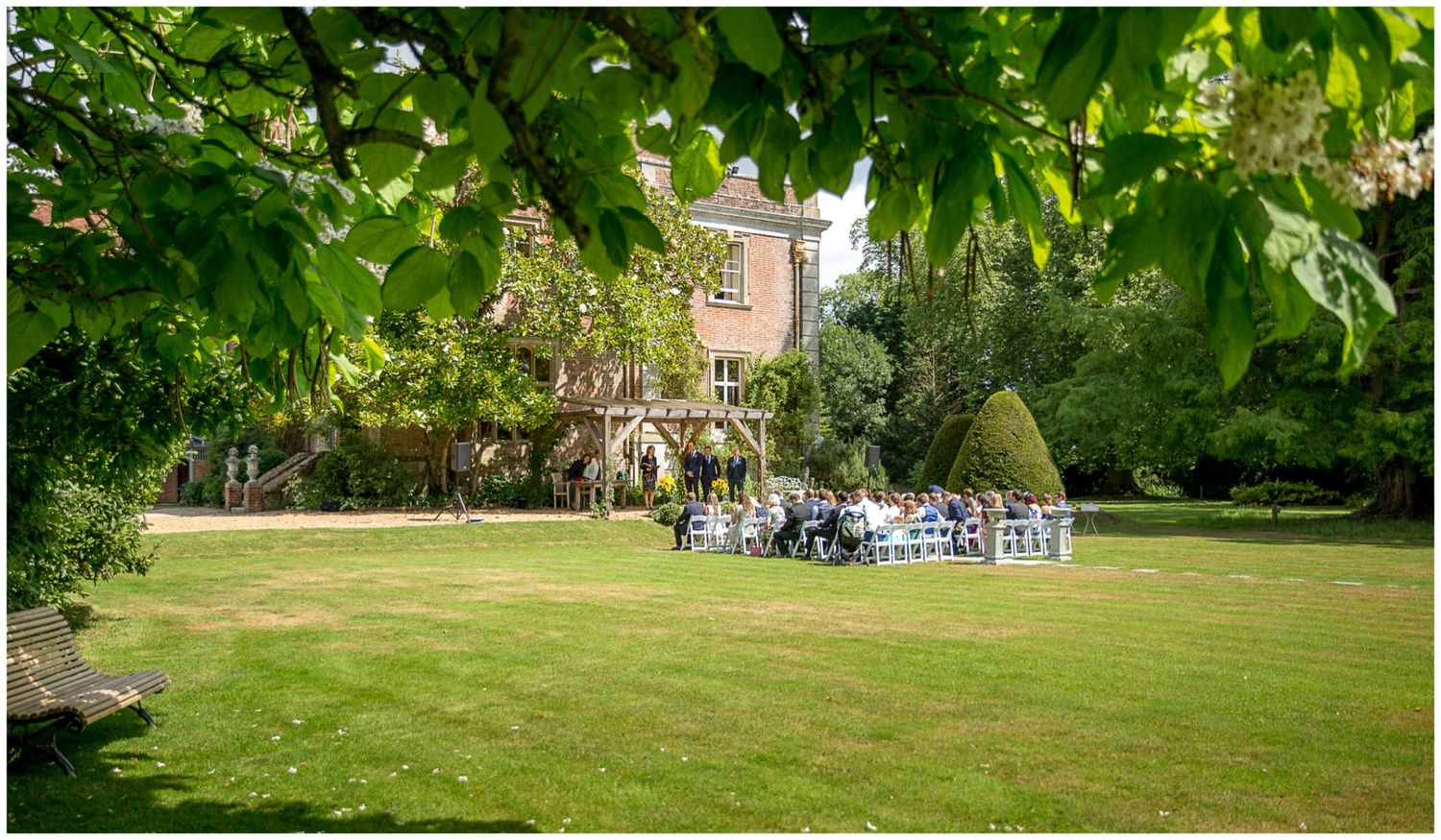 Wide angle view of a garden wedding venue with chairs arranged on manicured lawn surrounded by greenery.