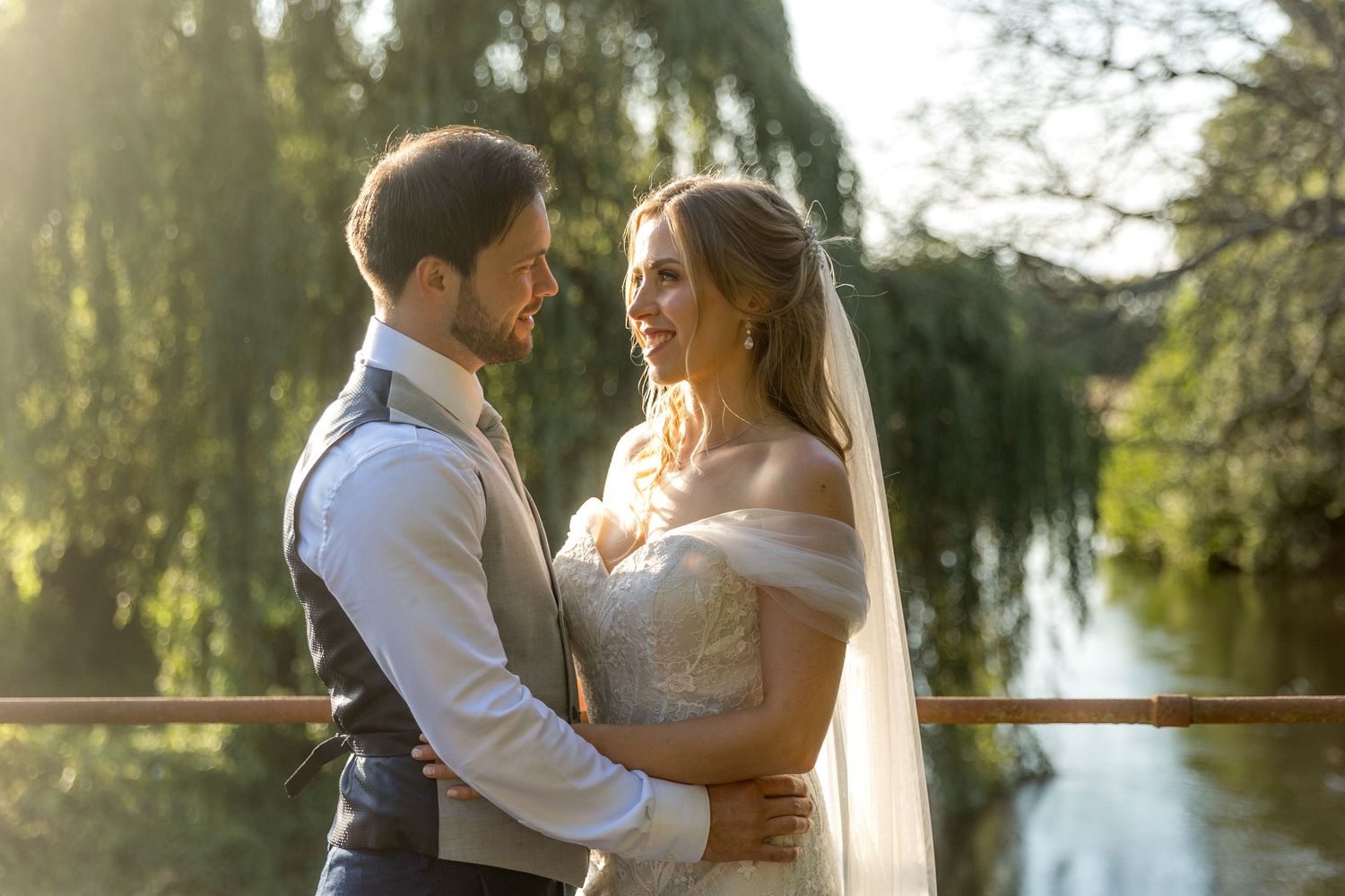 A romantic couple embraces during a golden hour photoshoot by a serene lake with willow trees in the background.