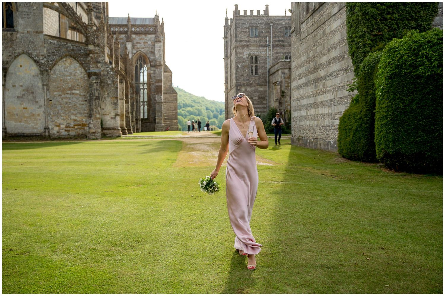 A person in a pink dress walks along the green lawn of a historic stone castle estate on a sunny day.