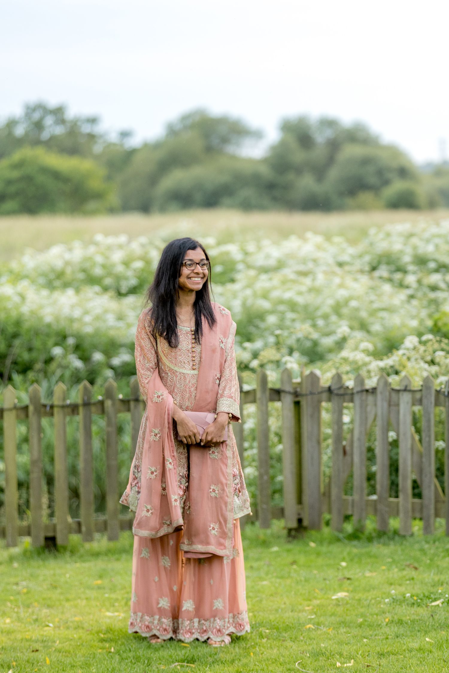 Woman in a flowing pink floral dress poses in a garden with white wildflowers and wooden fence.