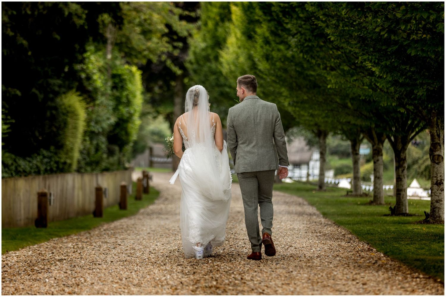 Long tree-lined pathway with wedding couple walking away from camera in natural outdoor setting.