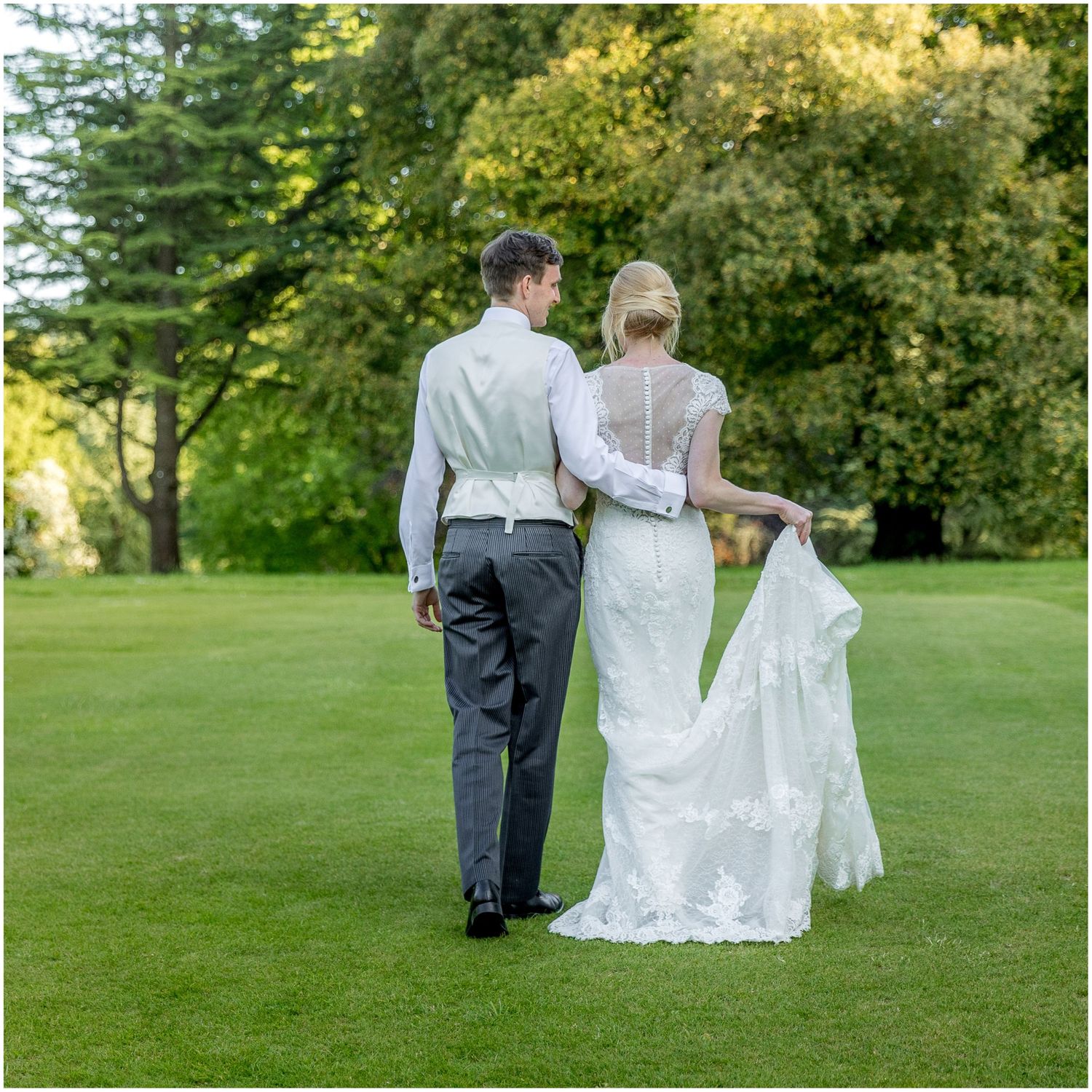 Newlyweds walking together on lush green lawn with trees in background during summer wedding.