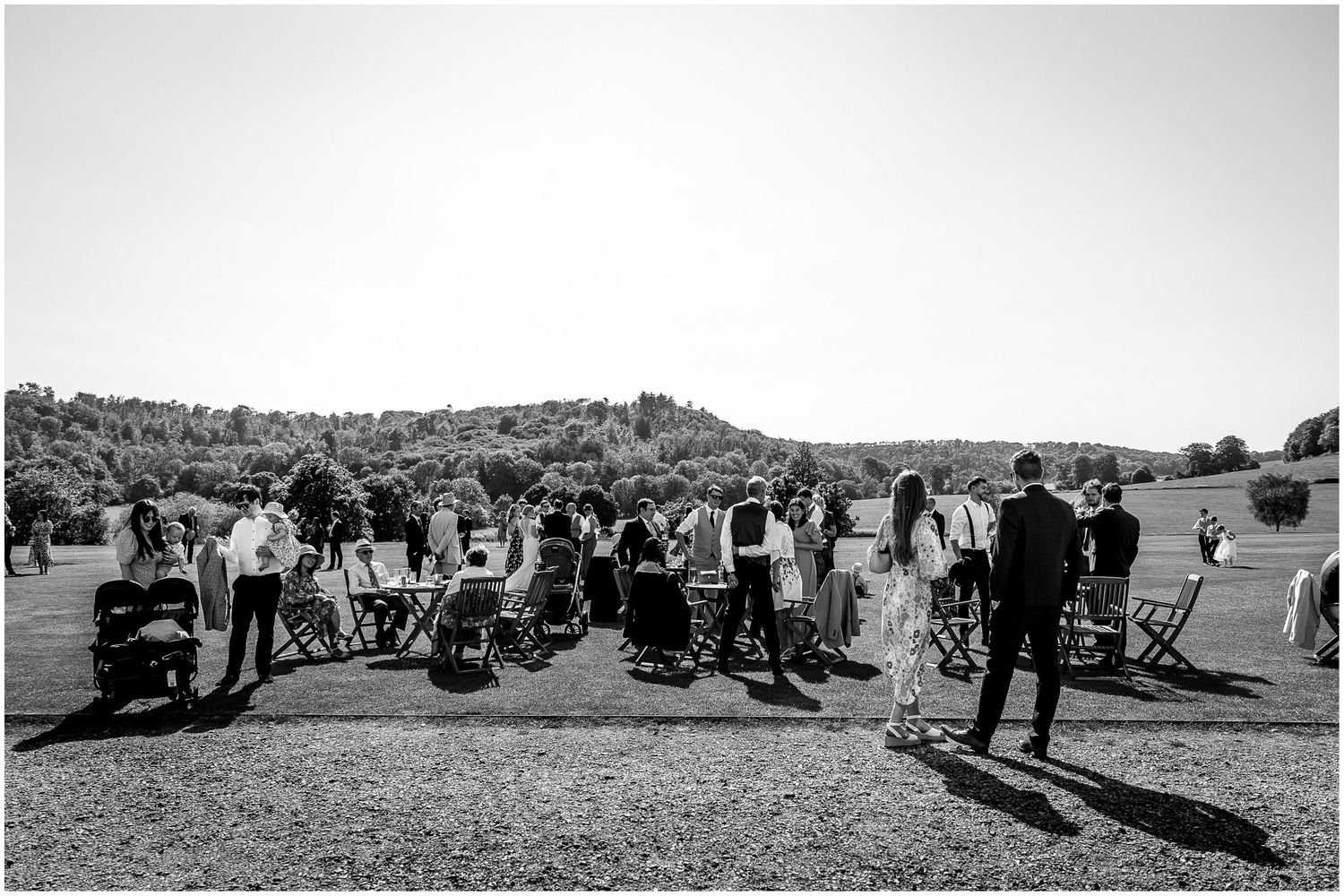 Black and white photo of wedding guests mingling on a hillside with chairs scattered around.
