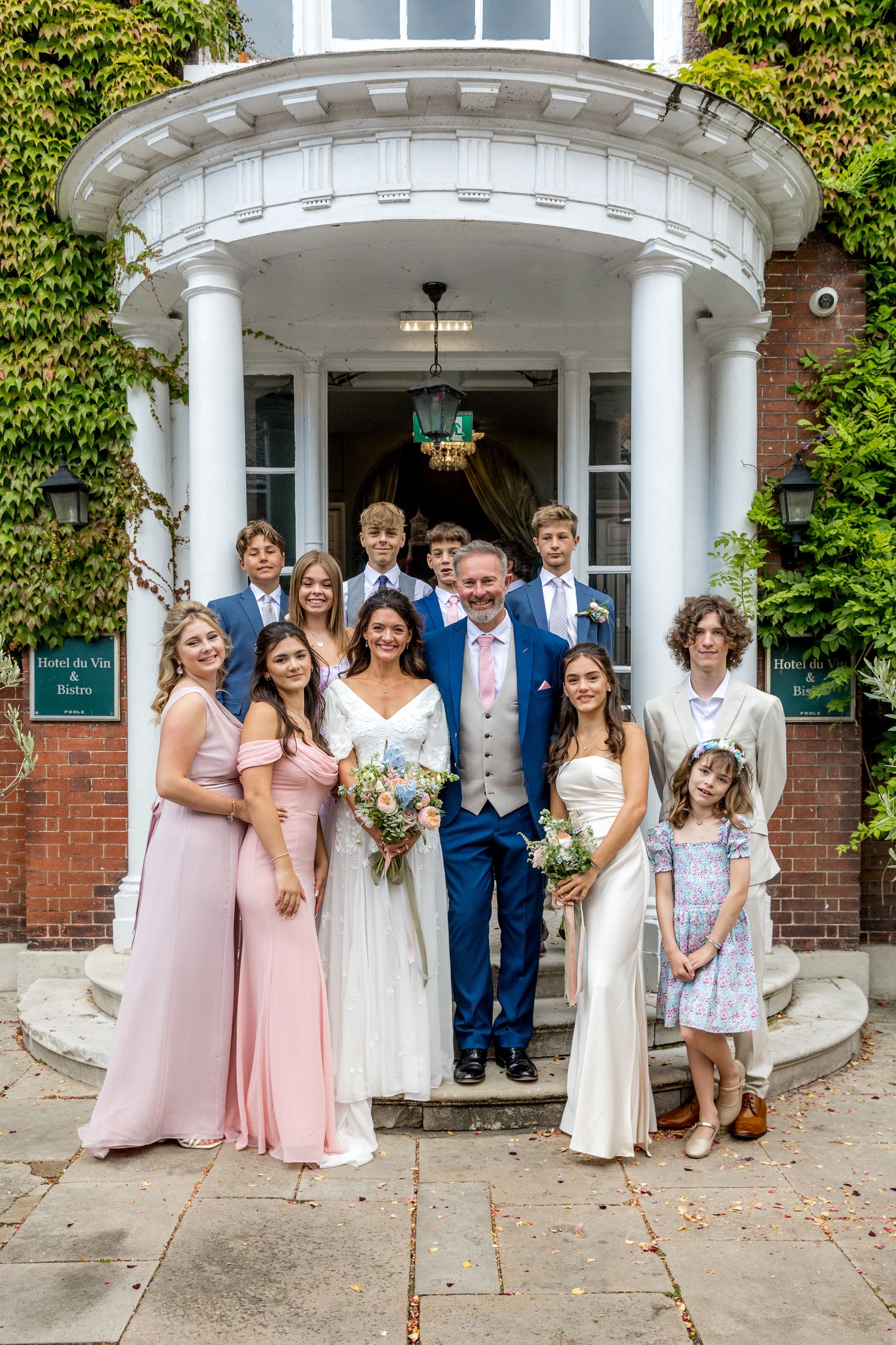 Wedding party poses in front of a beautiful brick building entrance with white columns and ivy covered walls.