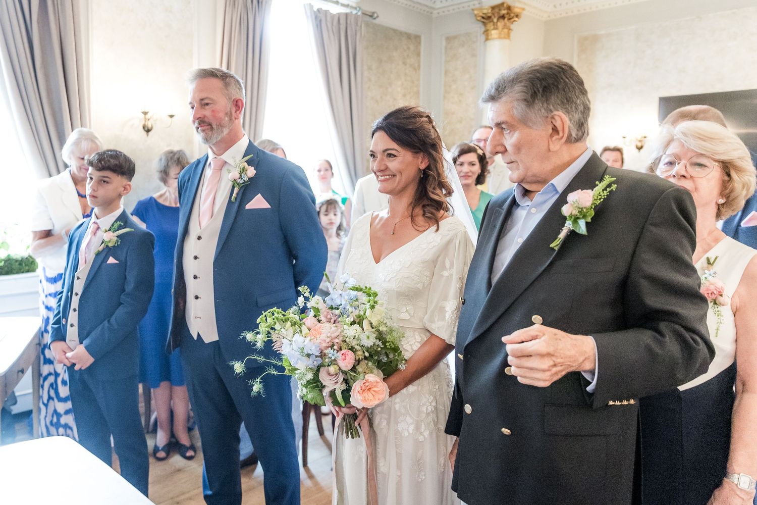 Wedding party gathered in an elegant room with natural light streaming through tall windows.