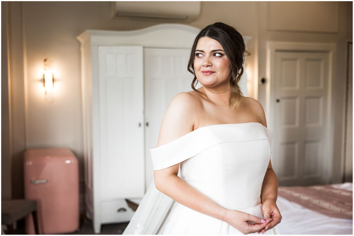 A bride in an elegant off-shoulder white wedding gown stands in a soft-lit room with classic decor and white doors.