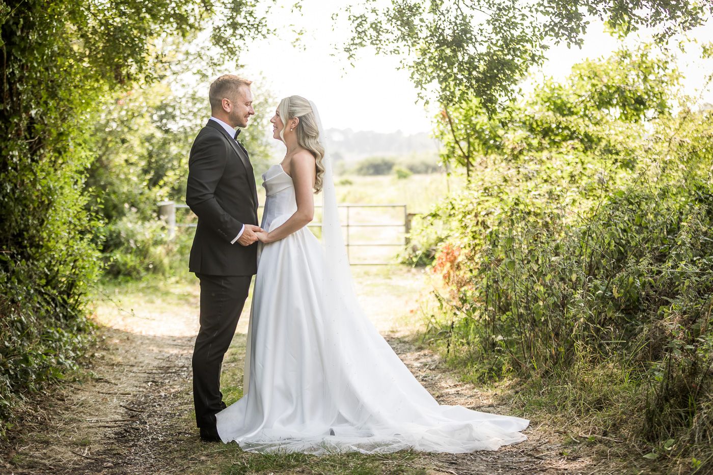 Romantic wedding couple poses together on a sunlit outdoor path surrounded by greenery.