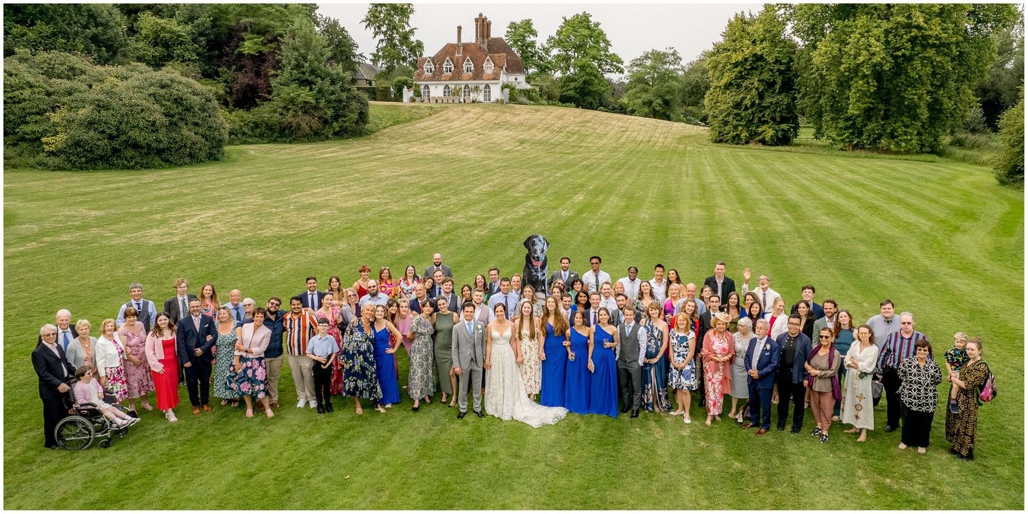 Large group wedding photo with guests gathered on a rolling green lawn.