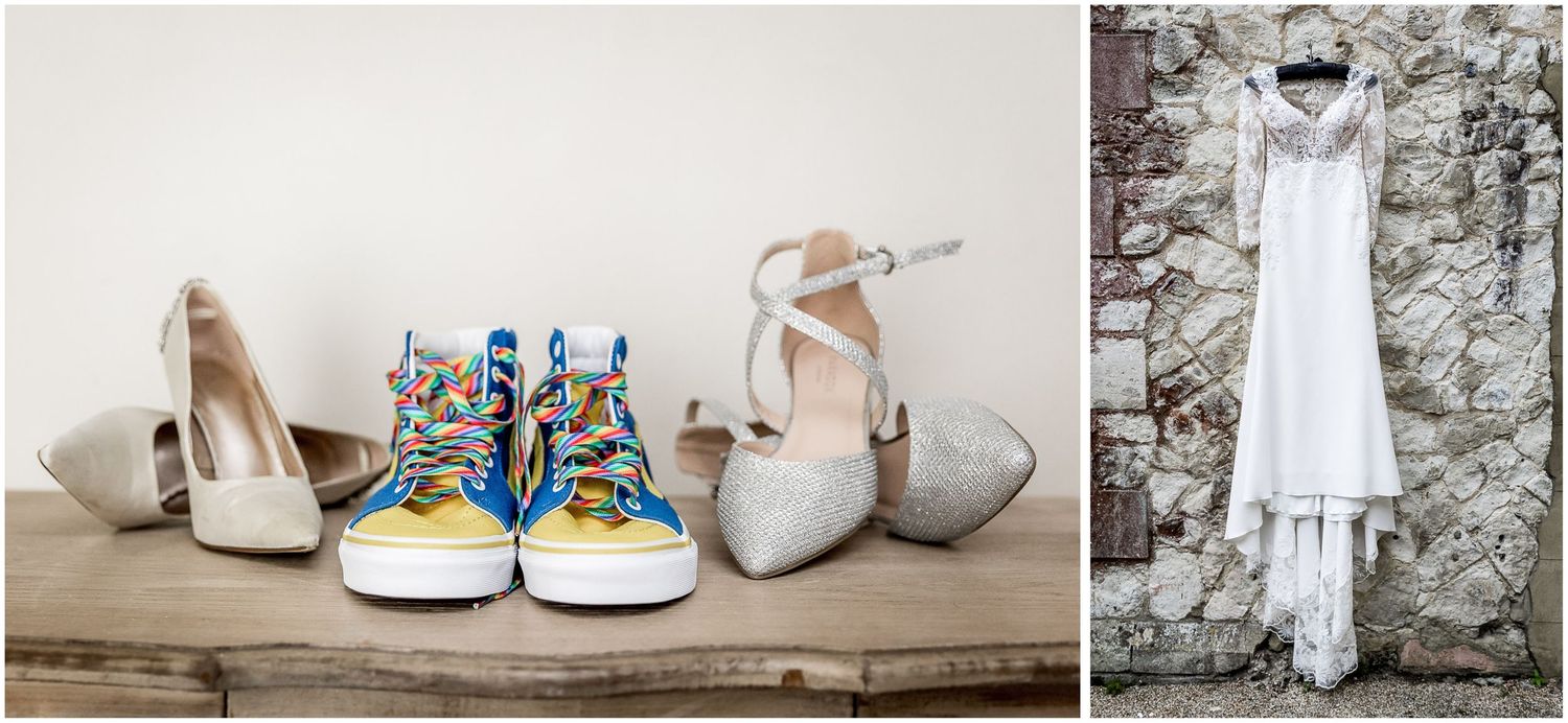 Wedding shoes including heels and colorful sneakers arranged next to a hanging white dress against stone wall.