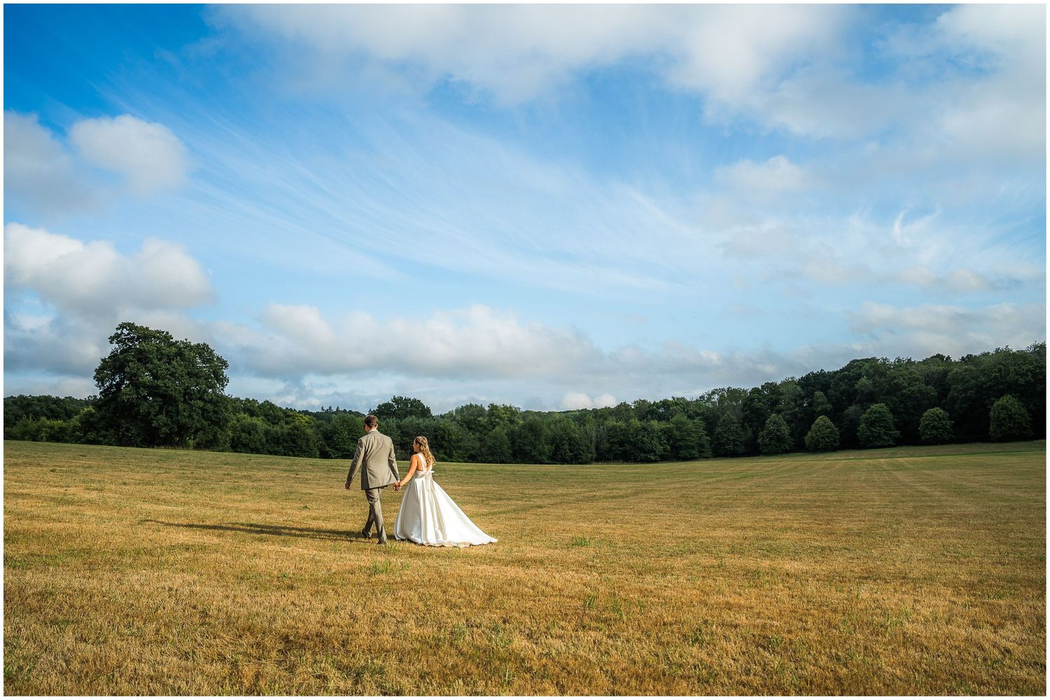 The wedding couple walk across a field bathed in evening sunshine as part of their wedding portrait photography session