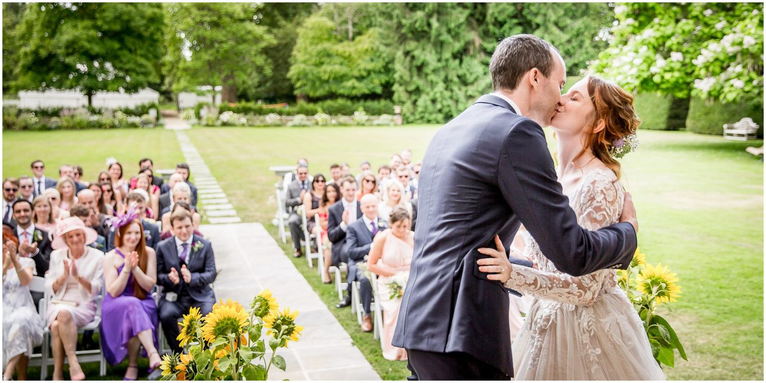 Romantic outdoor wedding ceremony on a lush green lawn with guests seated in rows watching the couple share their first kiss.