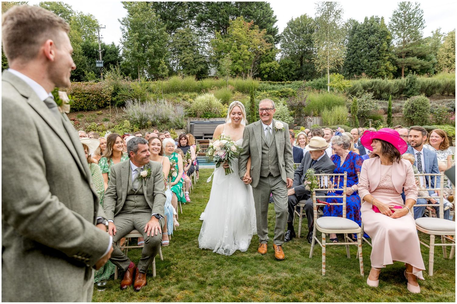 Panoramic view of outdoor wedding ceremony with guests seated on white chairs in a garden setting.