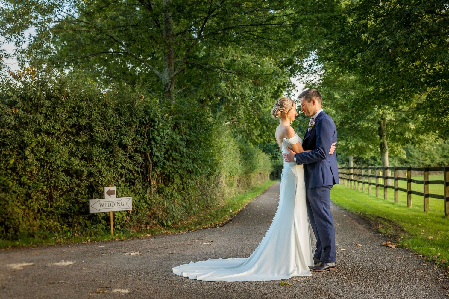 A couple strolls down a tree-lined path with wooden fencing on a sunny afternoon.