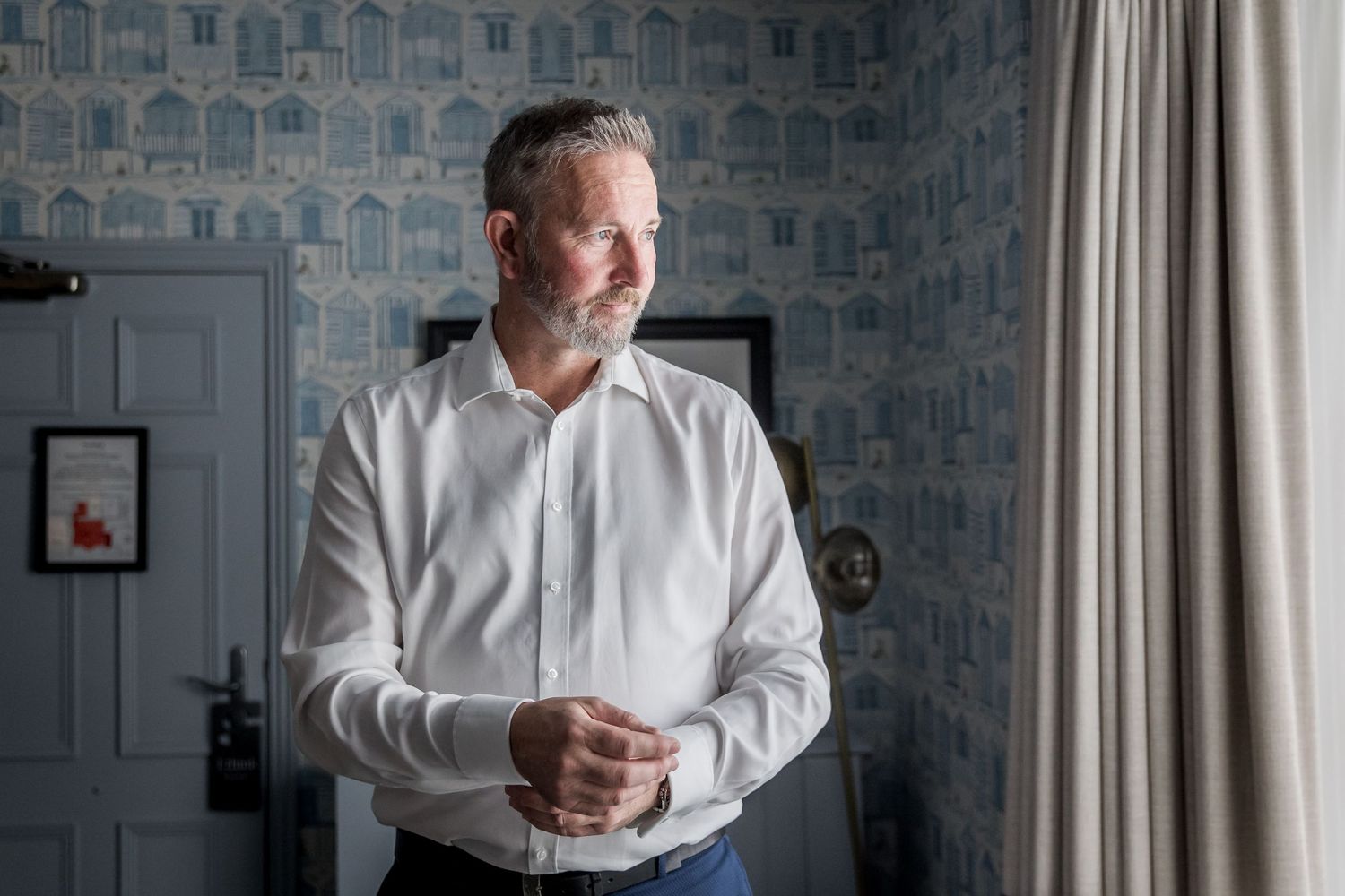 Man in white dress shirt photographed against patterned wallpaper.