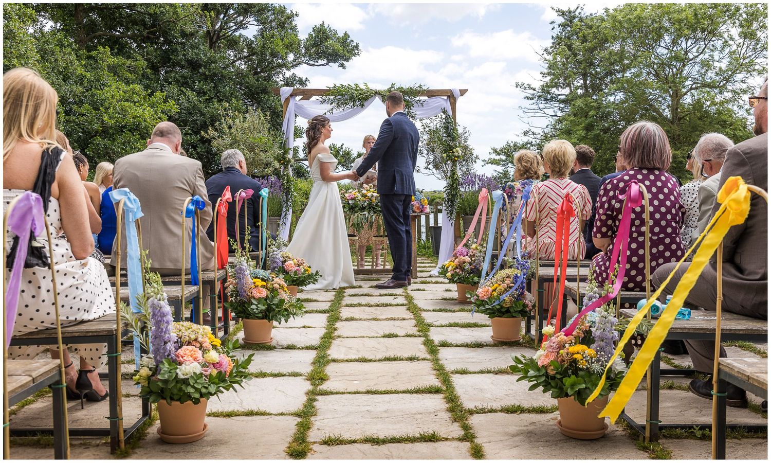 Sopley Mill's outdoor wedding ceremony space with colourful ribbon-decorated chairs lining a grass aisle leading to a floral arch.