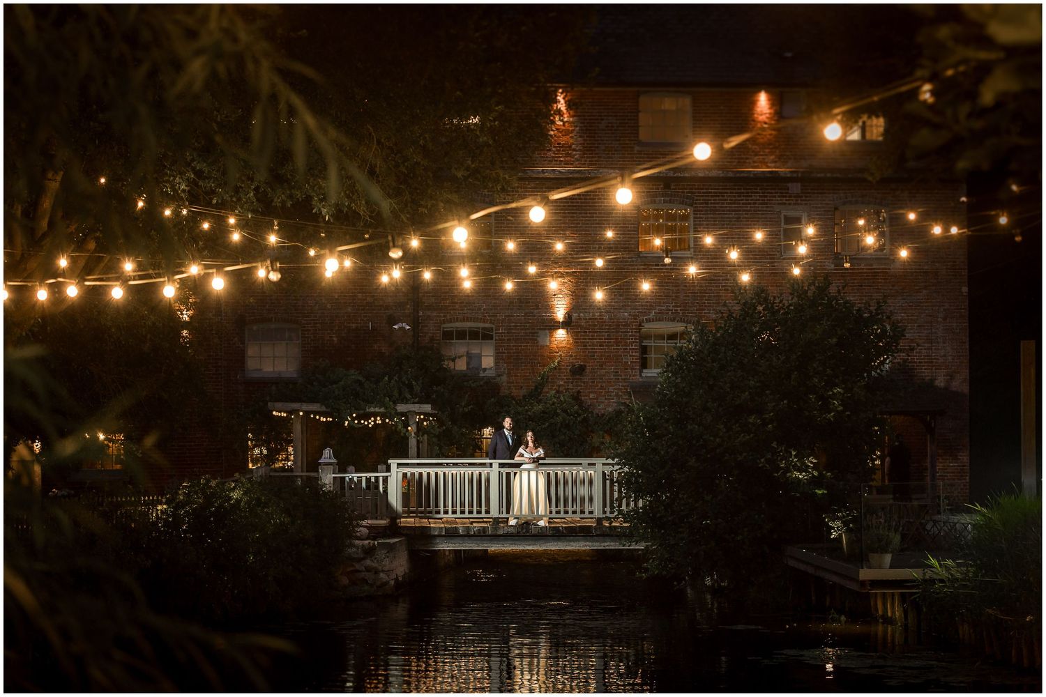 Couple on a romantic waterfront deck at night, surrounded by glowing string lights and lush greenery.