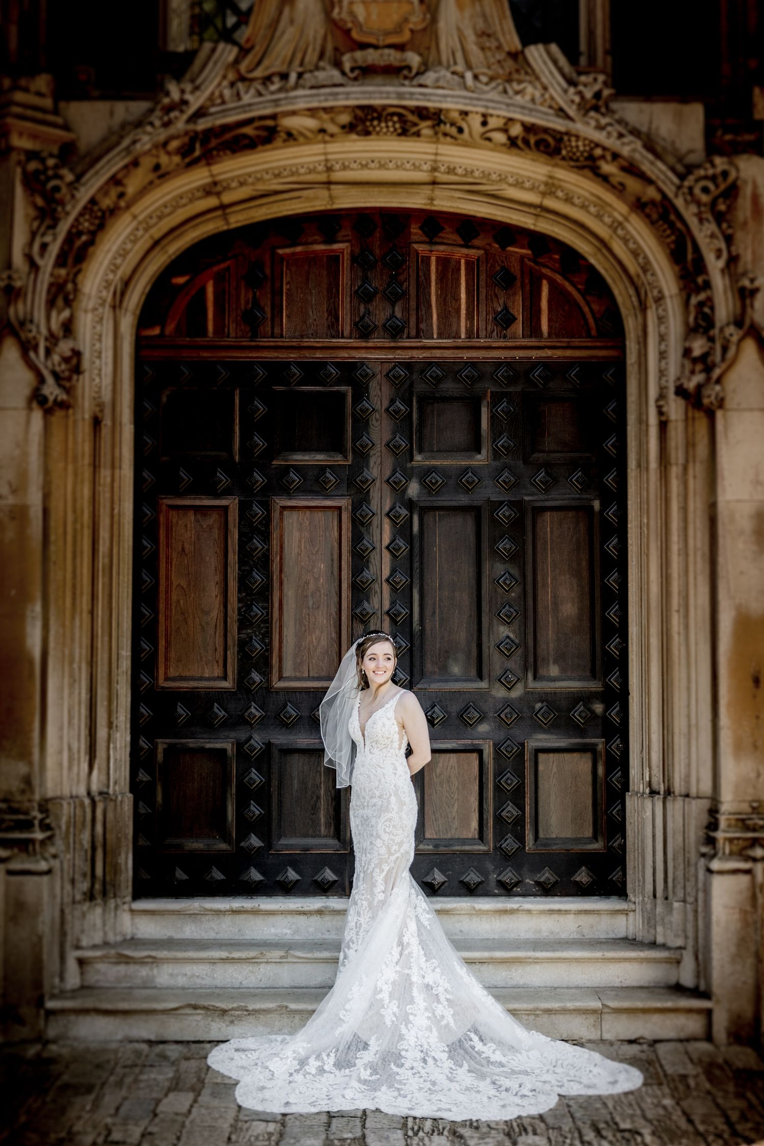 A bride in a white lace gown with long train poses in front of ornate gothic doorways and stone archways.
