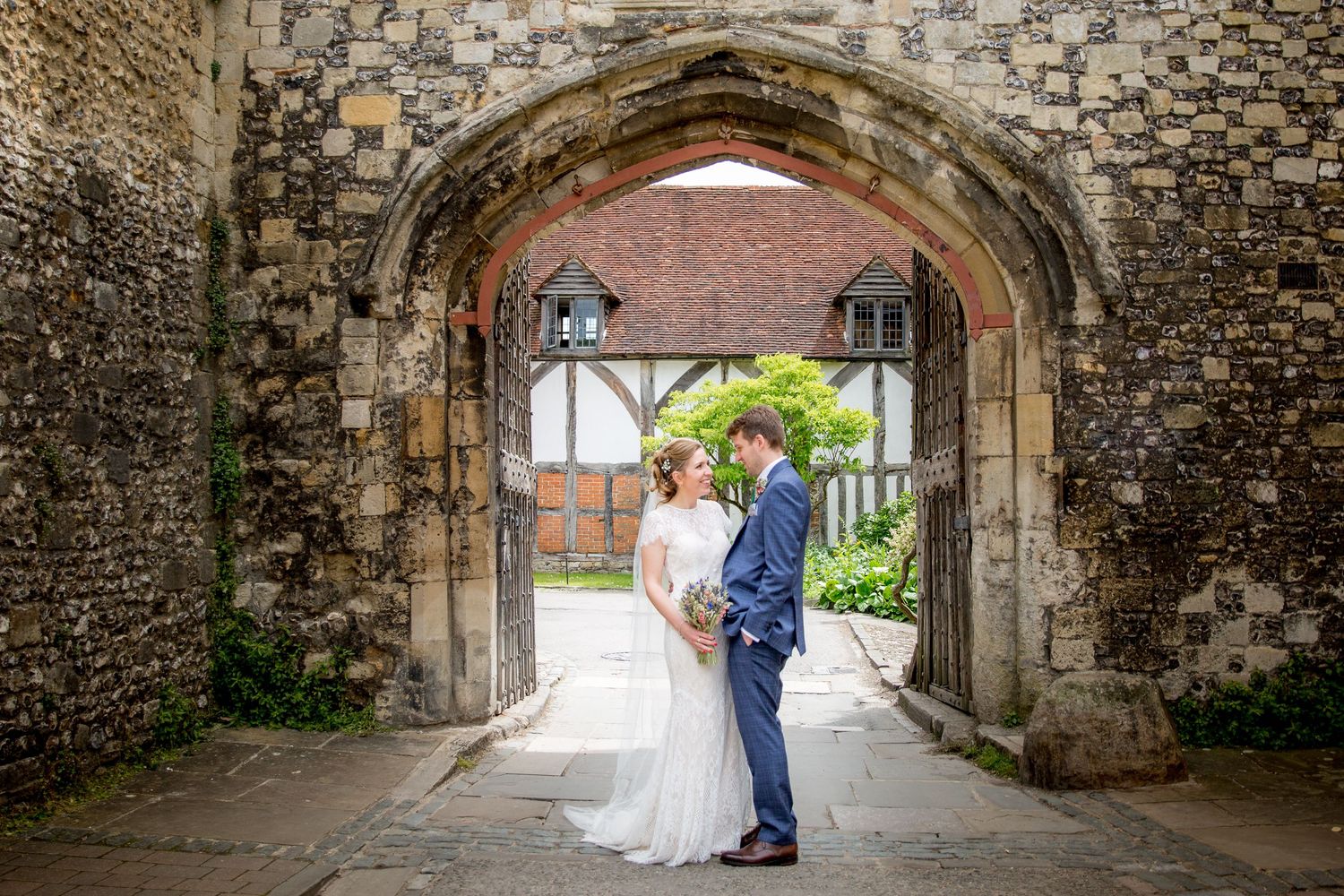 A couple in wedding attire stands beneath a stone archway at a historic venue with lush greenery visible beyond.
