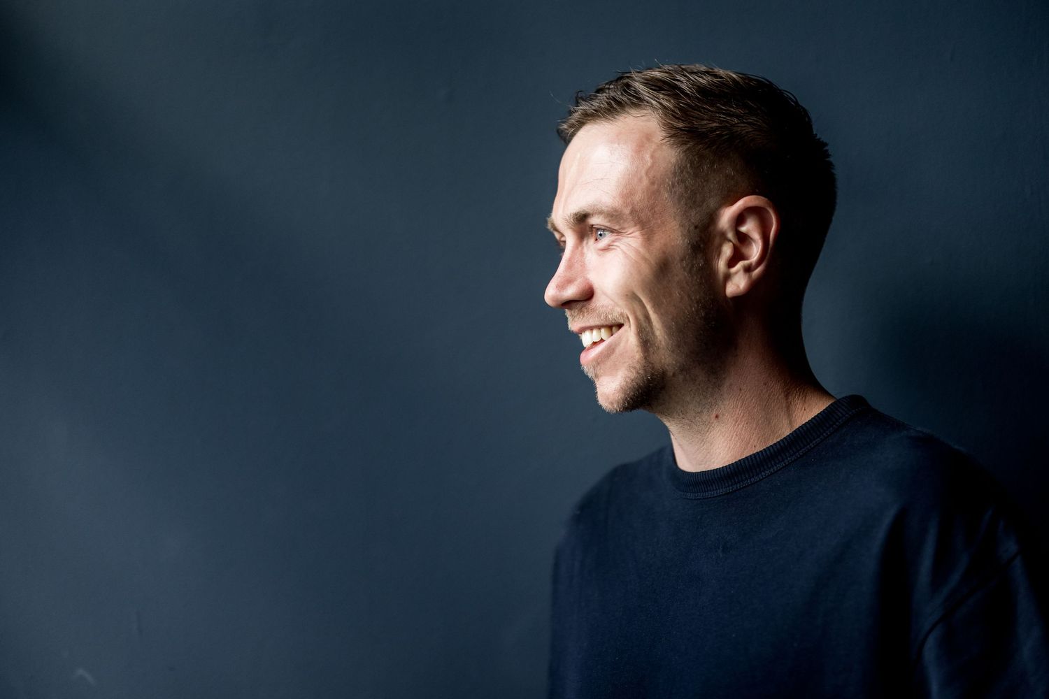 Professional studio portrait of person in navy sweater against dark background with dramatic side lighting.
