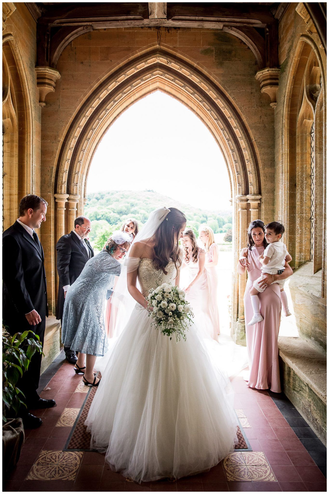 Wedding party gathered under elegant stone archways in a historic venue with natural lighting.