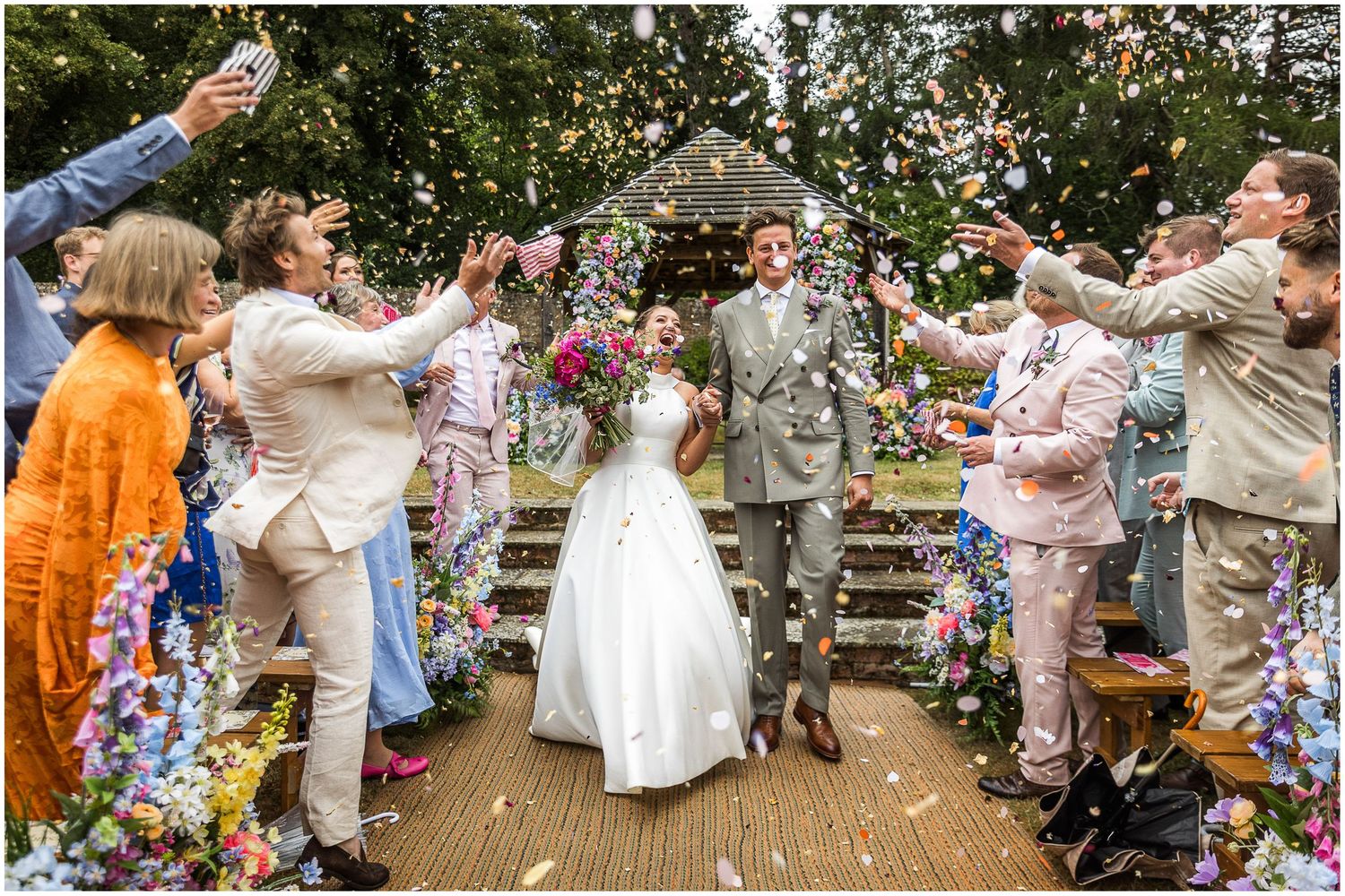 The newly married bride and groom walk down the aisle together showered by confetti from guests at the outdoor ceremony in the walled garden