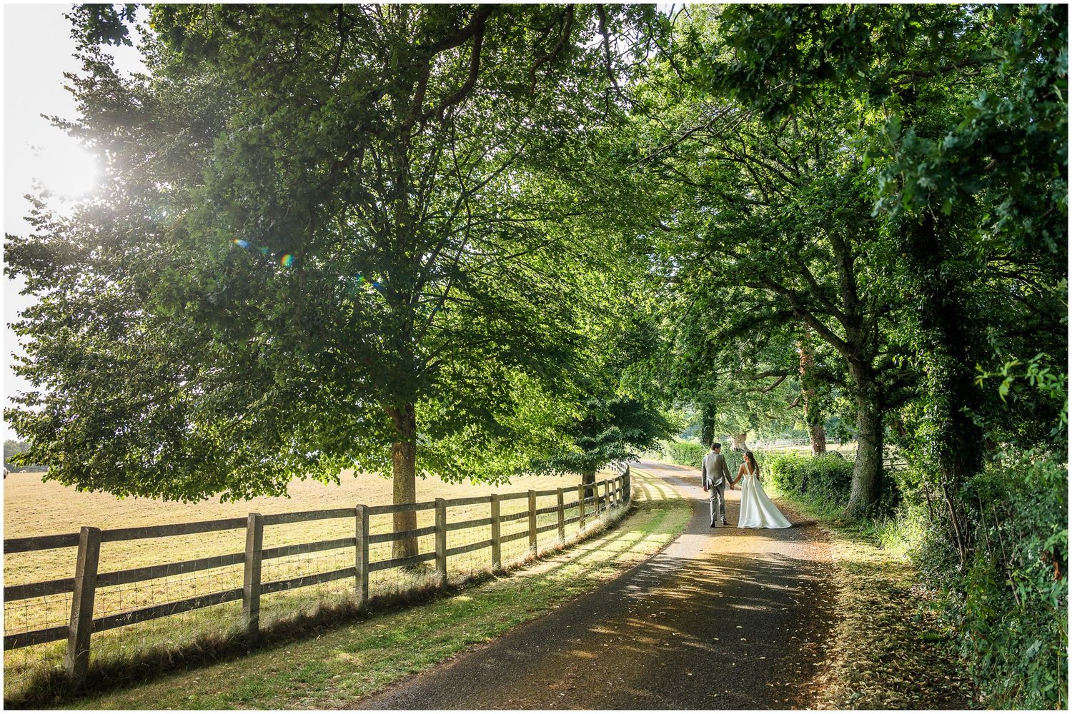 Bride and groom walking along a tree-lined drive with wooden fence, bathed in soft sunlight at the Holywell Estate wedding venue