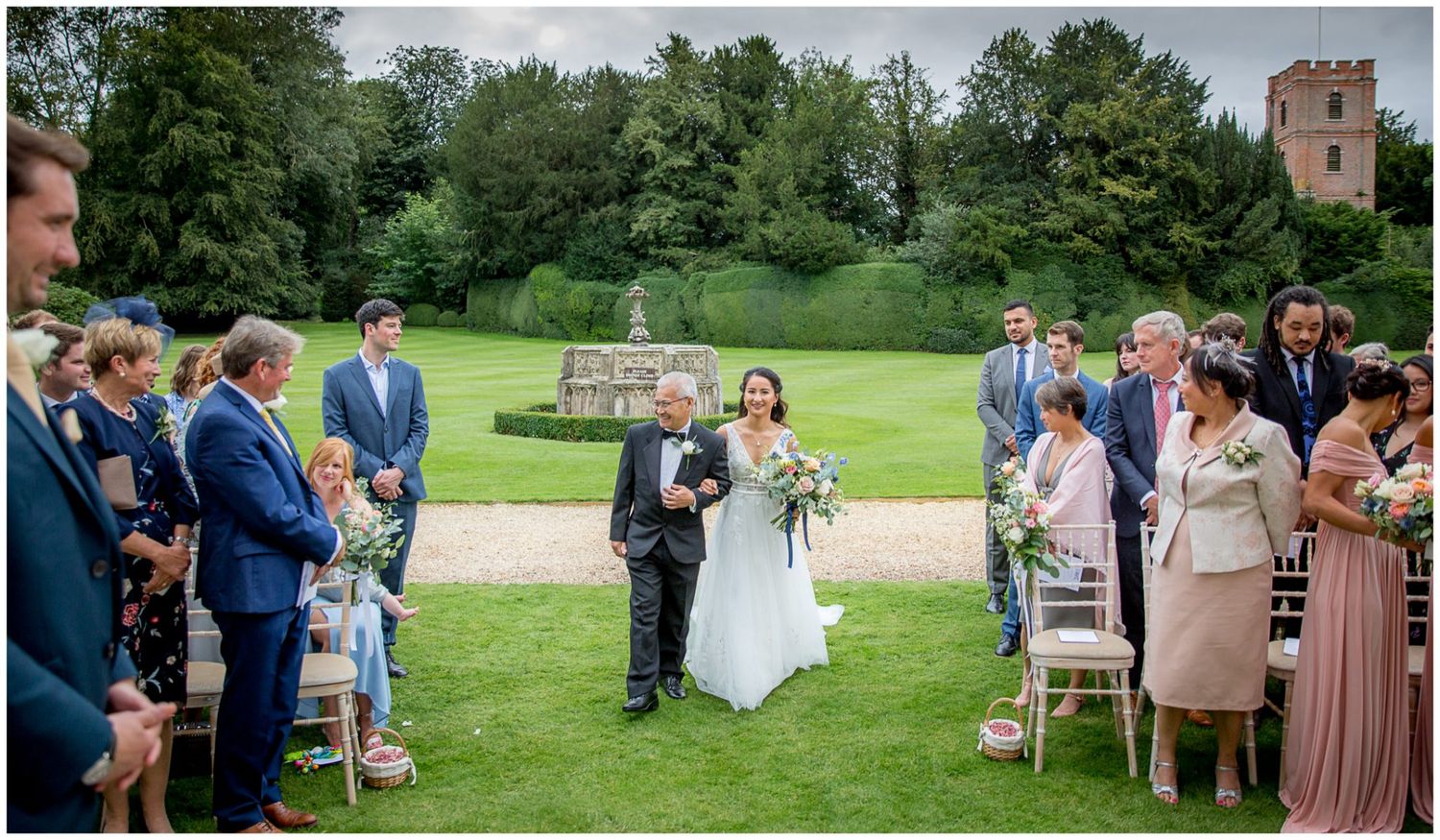 Wedding ceremony guests seated on white chairs on a lush green lawn with historic architecture in the background.