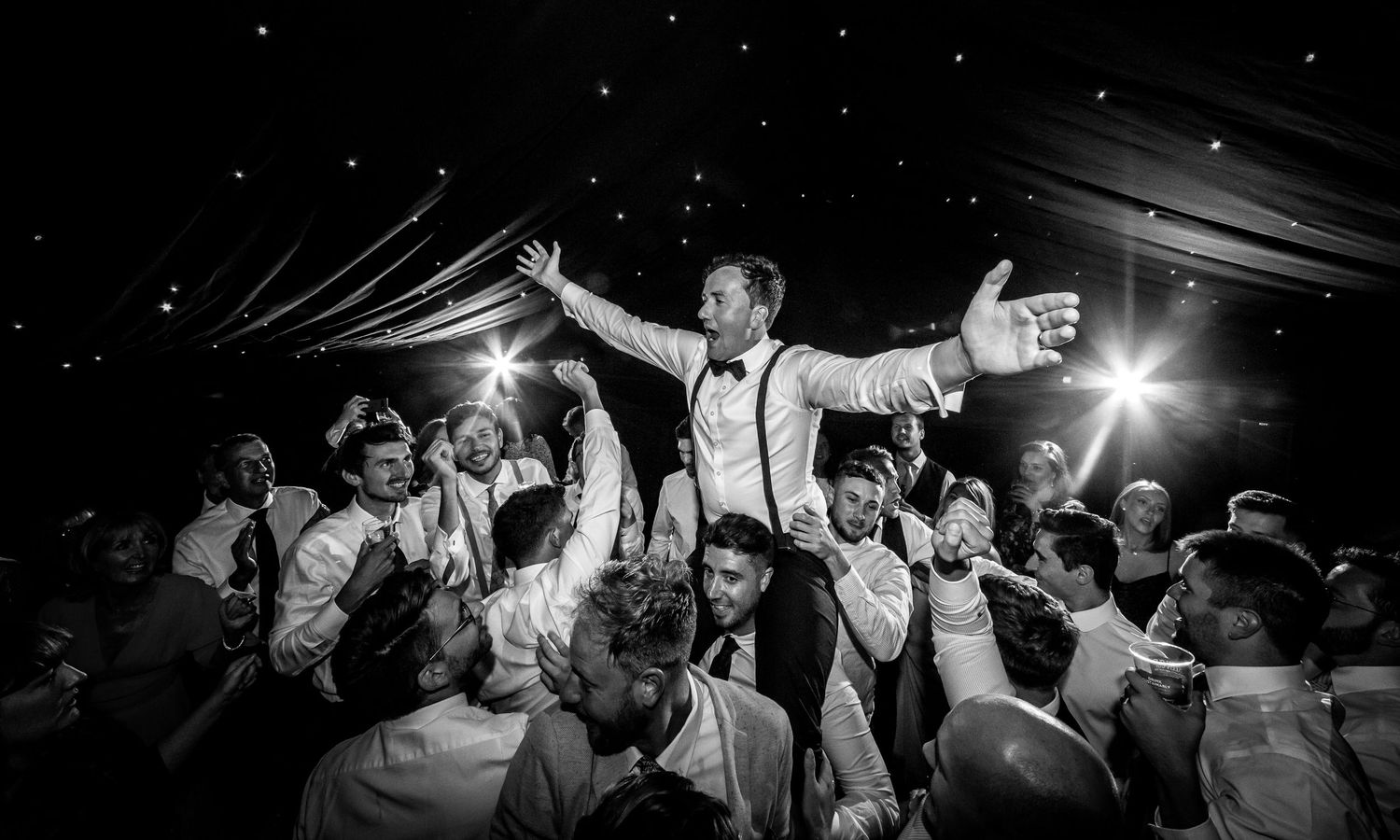 Wedding party dancing and celebrating under dimly lit reception tent at night.
