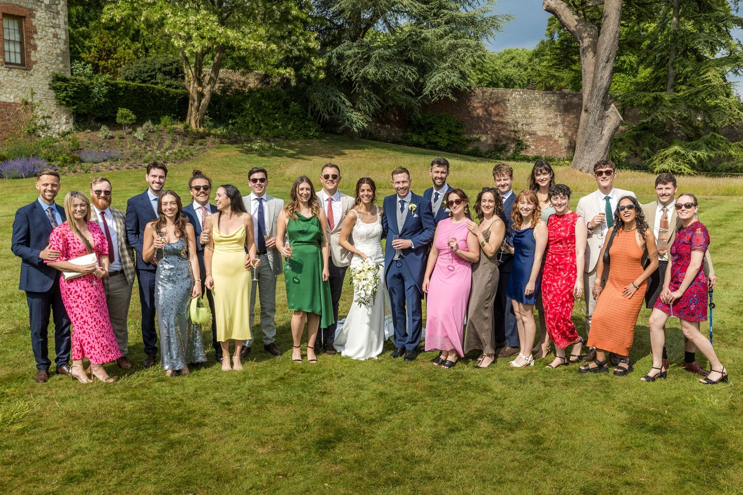 Large wedding party group photo with colorful dresses arranged on grassy outdoor venue.