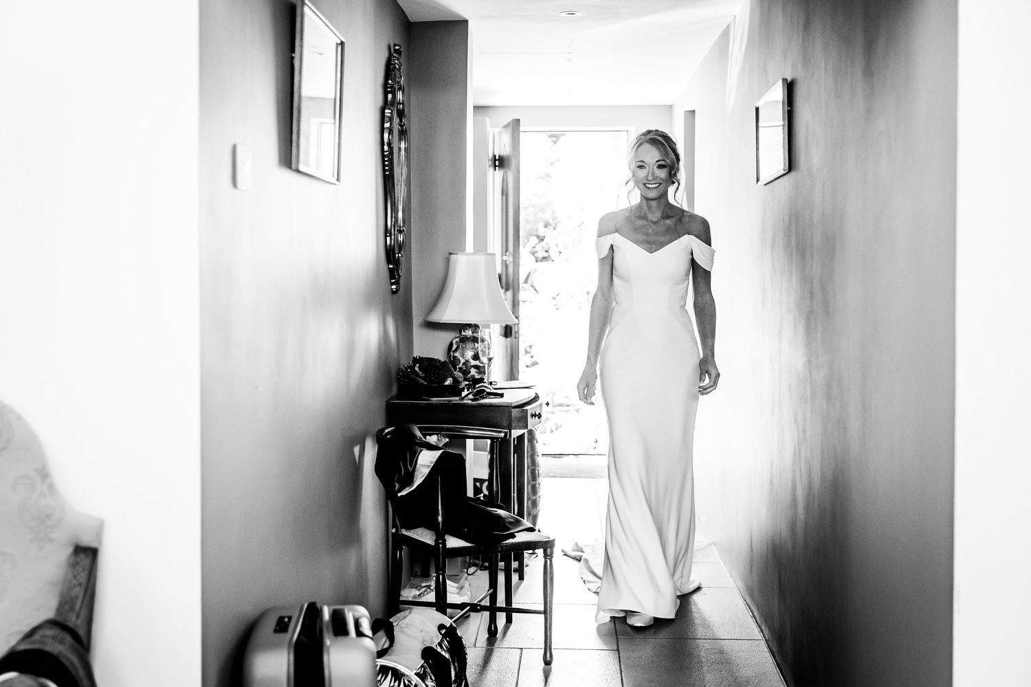 A bride in a sleek white dress walks down a narrow hallway in black and white.
