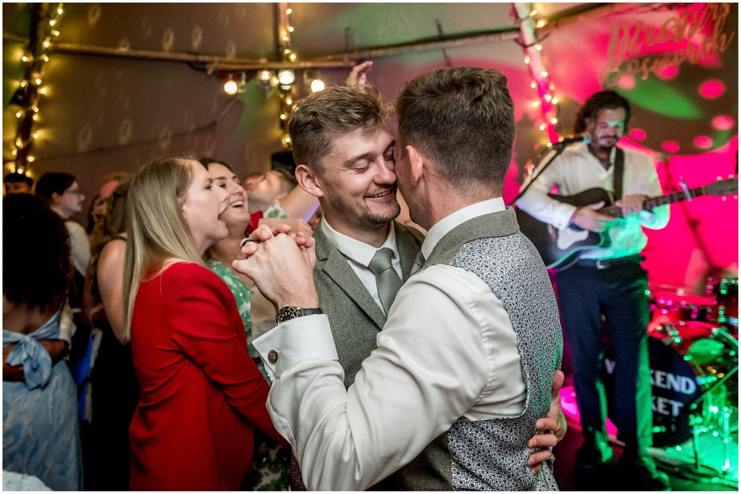 Wedding party celebrates on dance floor with colorful lighting and festive atmosphere.
