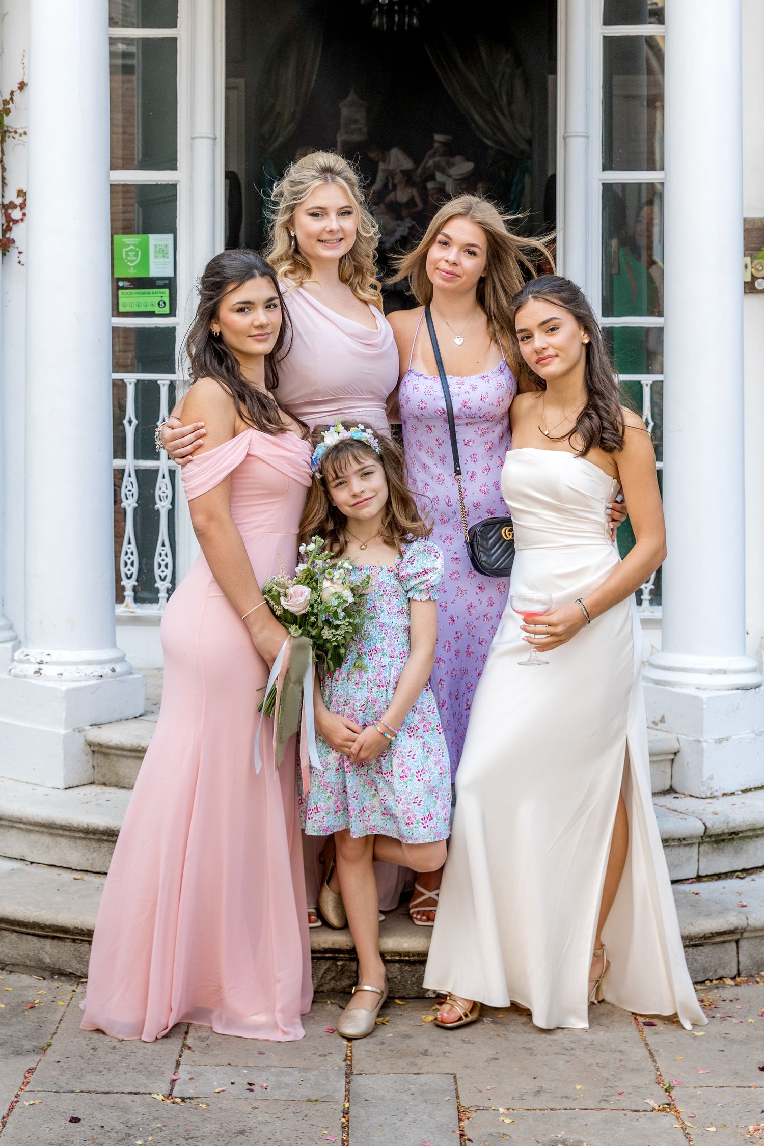 Group photo of wedding attendees in pink dresses gathered on stone steps near white columns.