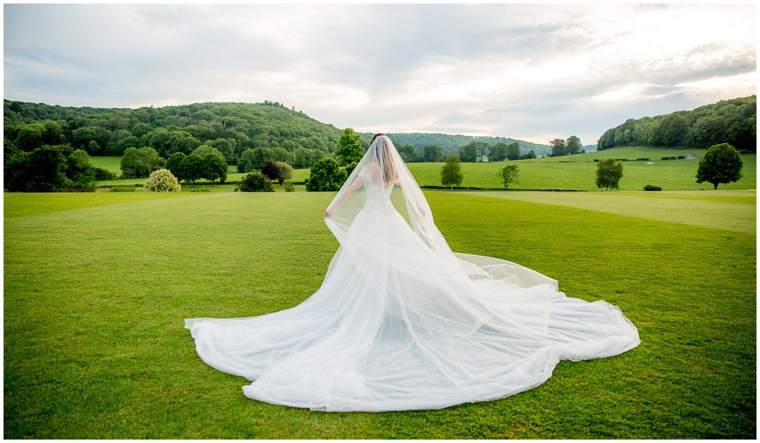 Long flowing white wedding dress train spread out on lush green grass with hills in the background.