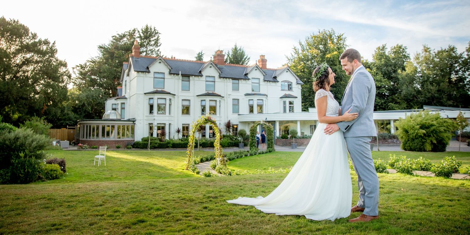 Couple poses on lawn in front of grand white Victorian mansion wedding venue.