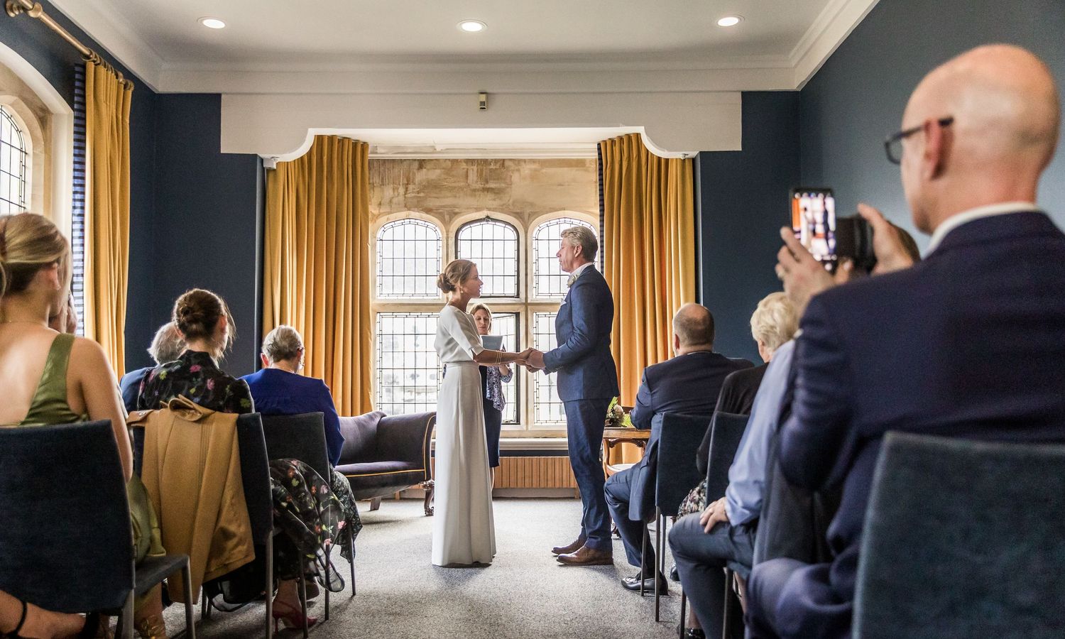 An intimate wedding ceremony takes place in an elegant room with gold curtains and arched windows.