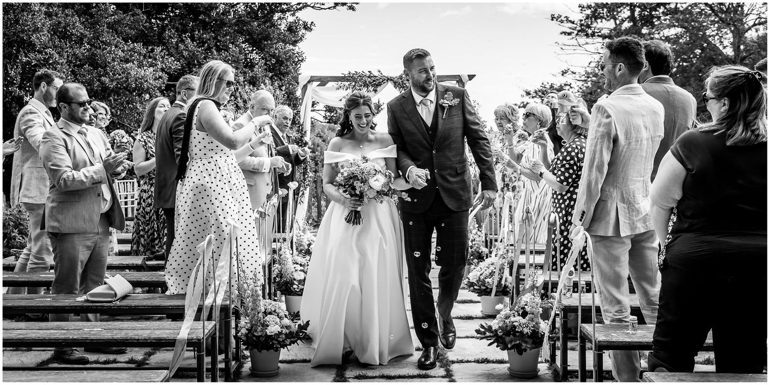 Black and white wedding photograph of a bride and groom walking down the outdoor aisle at Sopley Mill in the New Forest.