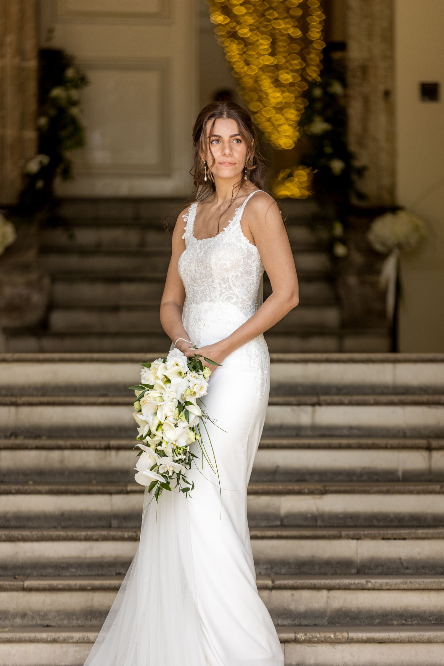 Elegant bride in white dress poses with bouquet on grand stone steps at outdoor venue.