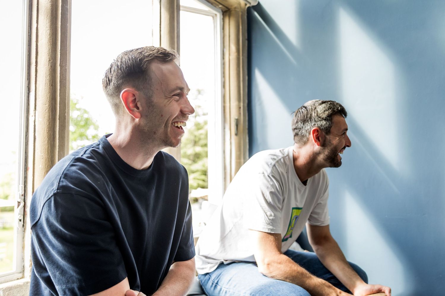 Two people sitting by a window sharing a relaxed conversation in a bright room.
