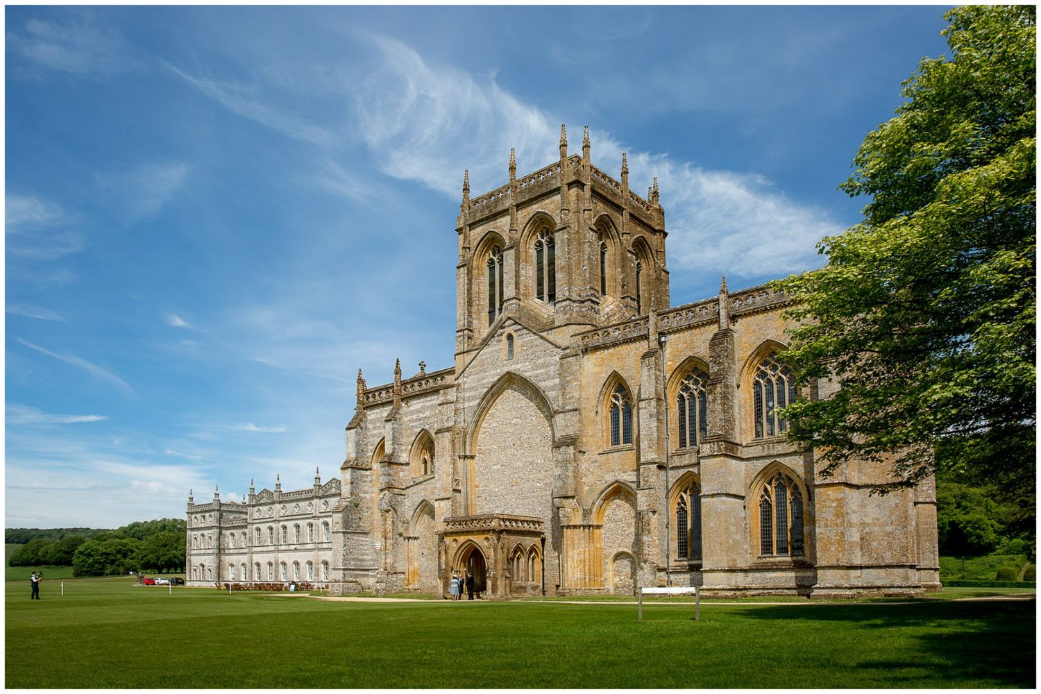 Historic Sherborne Abbey in Dorset stands majestically against a bright blue summer sky with green trees surrounding it.