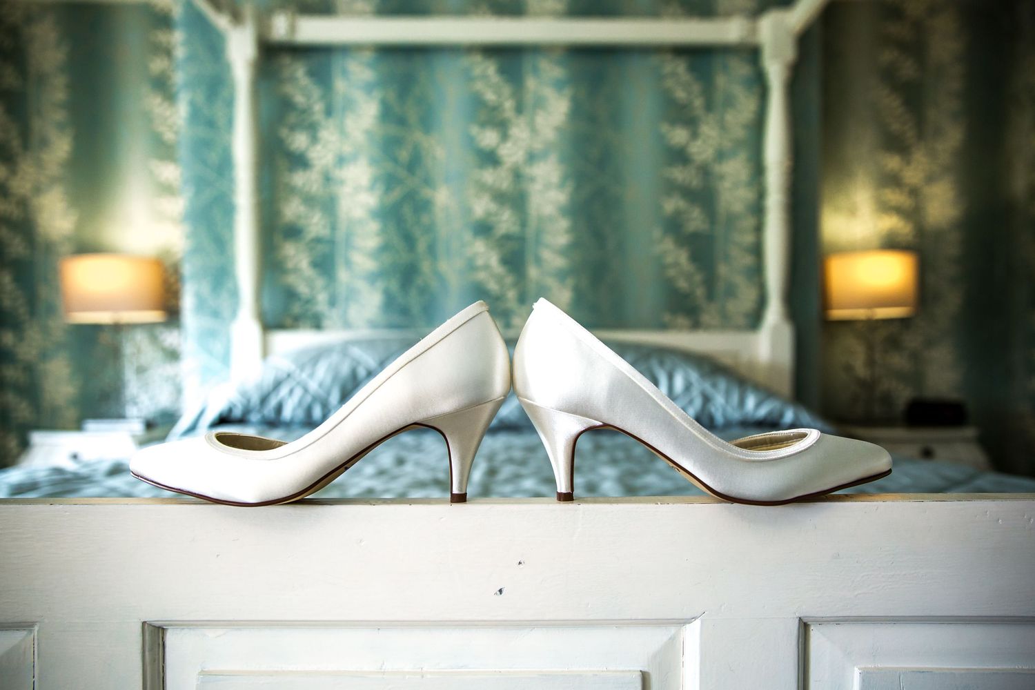 Row of elegant high-heeled bridal shoes displayed on a white shelf against turquoise wallpaper.