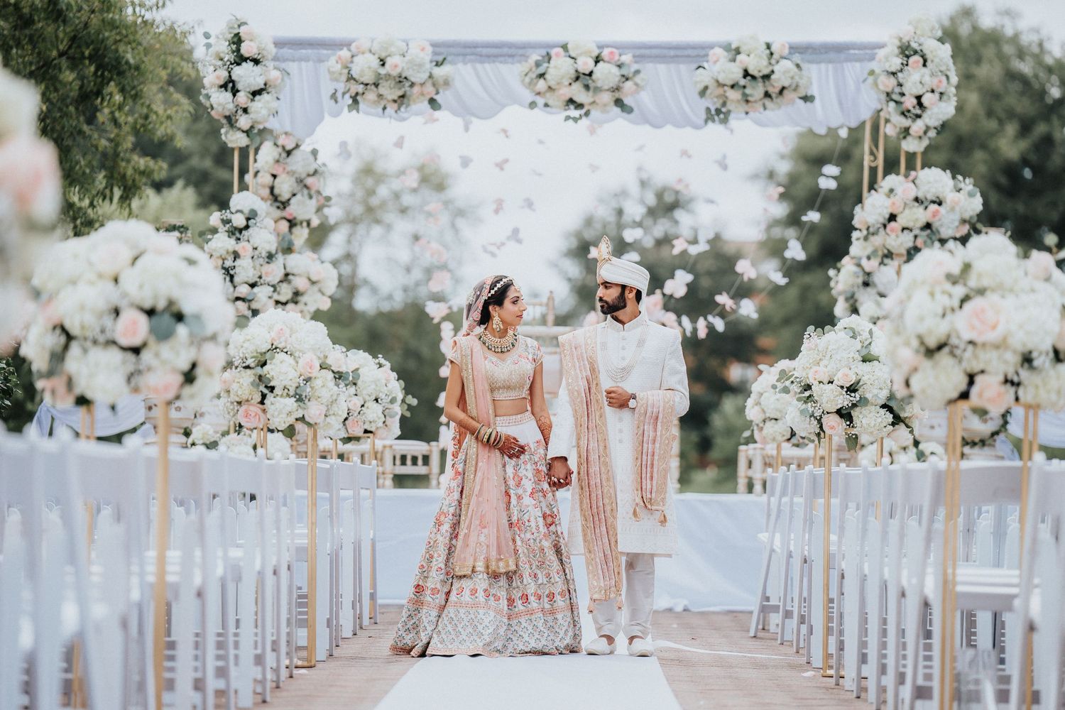 White floral arrangements line an outdoor wedding aisle with chairs and a waterfront backdrop.