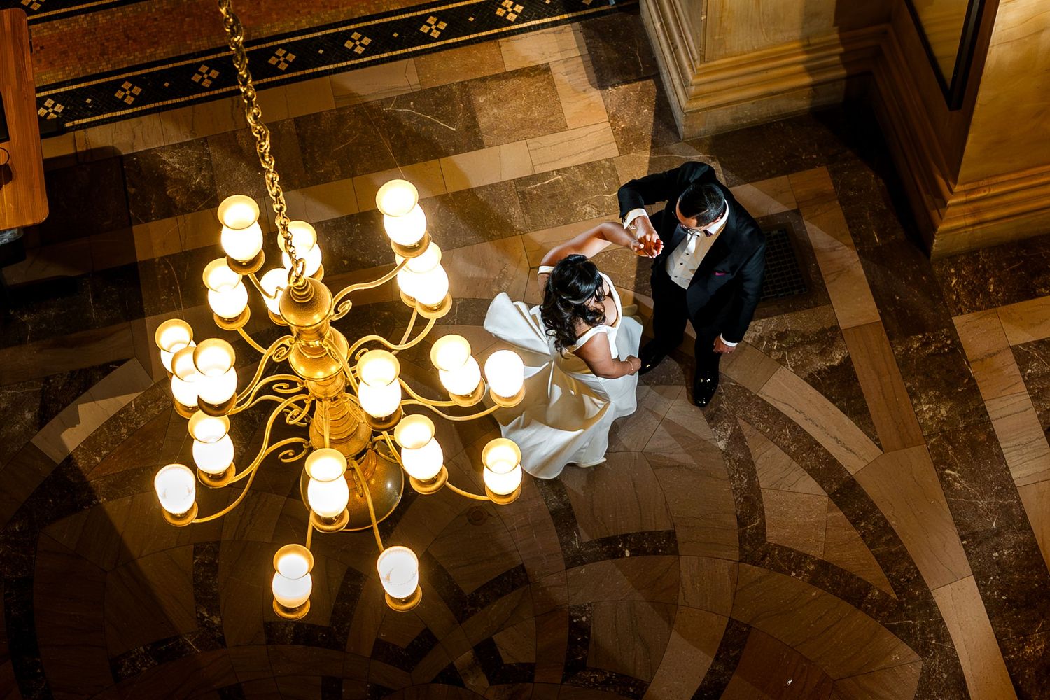 A couple dances beneath an elegant crystal chandelier in a grand ballroom with ornate marble flooring.
