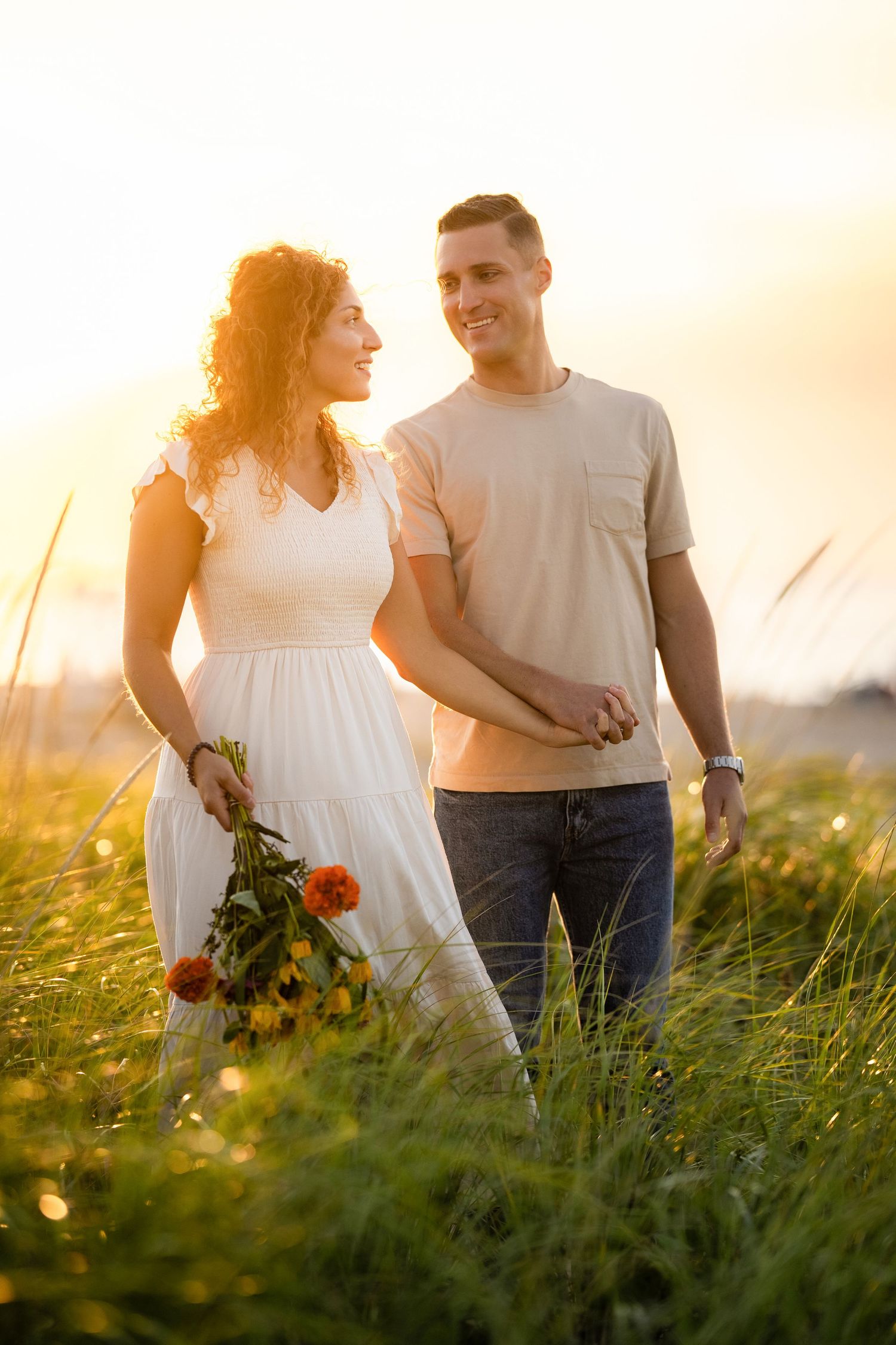 Couple walking together in a sunlit meadow at golden hour holding flowers and enjoying a romantic moment.