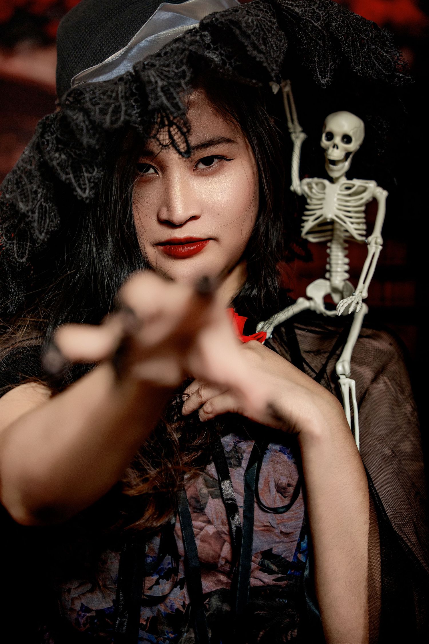 Gothic portrait with skeleton prop and black lace hat against dark background.