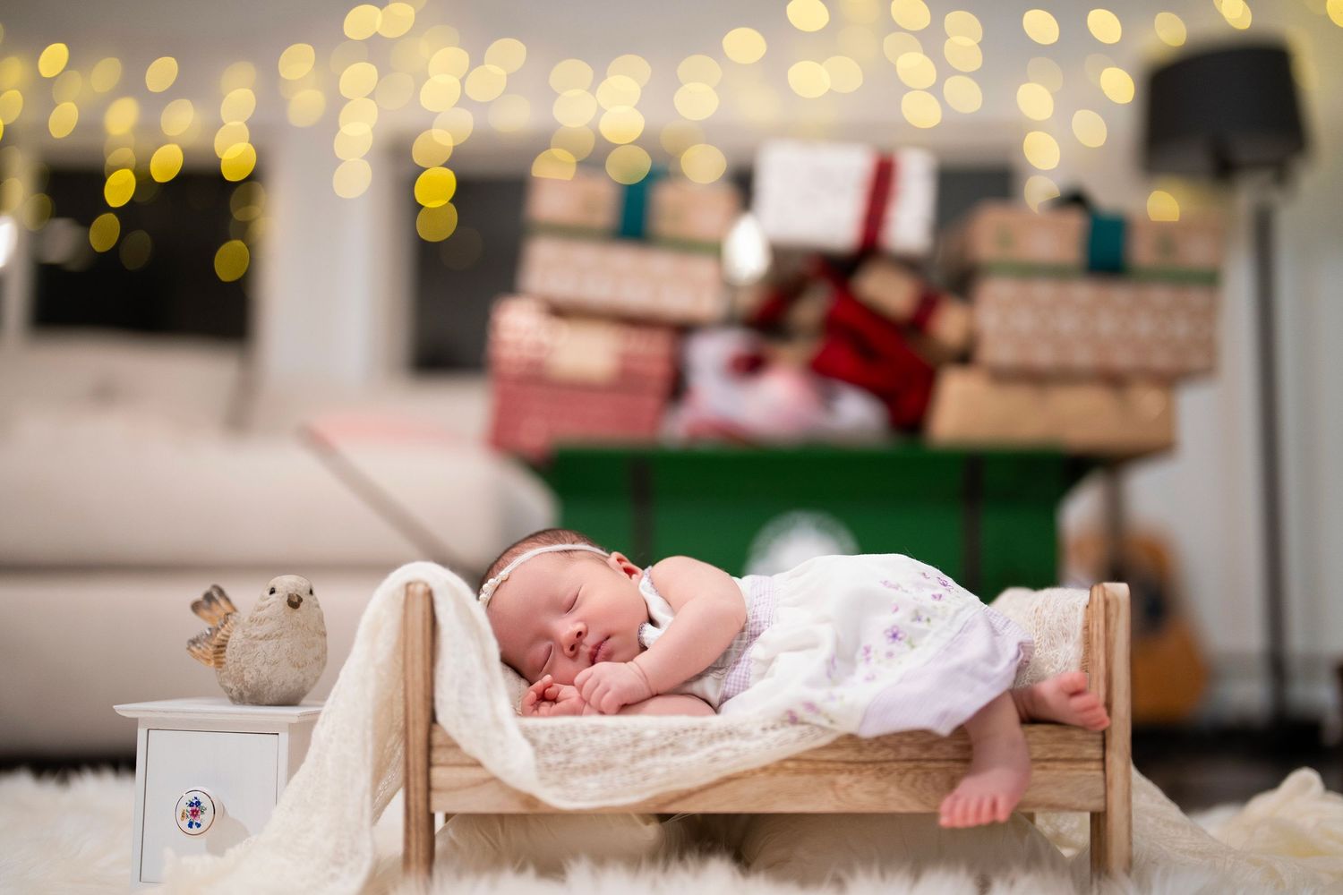 A sleeping newborn on tiny wooden bed with Christmas lights and presents in background.