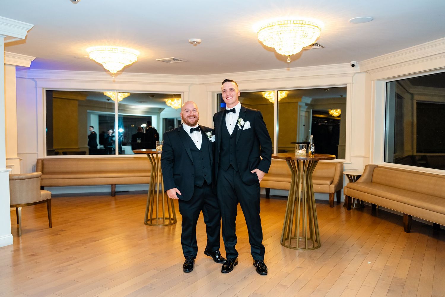 Two people in formal black tuxedos pose in elegant ballroom with chandeliers.