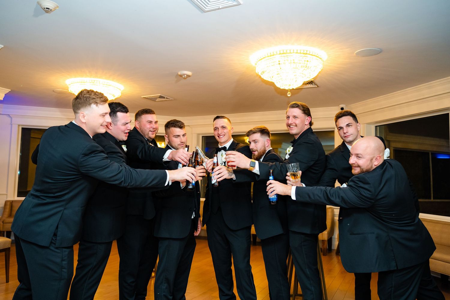 Groomsmen in black suits striking dramatic poses in an indoor venue with crystal chandeliers.