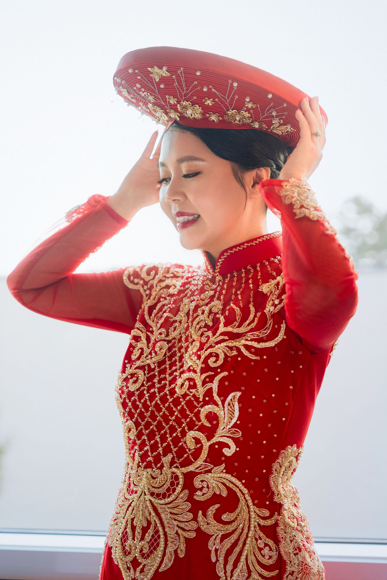Traditional Vietnamese bride wearing ornate red ao dai and circular headdress in sunlit setting.